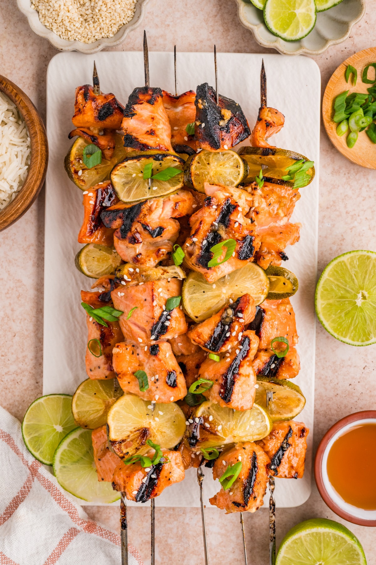 Grilled honey soy lime salmon skewers on a serving platter with charred lime slices, sesame seeds, and green onions.