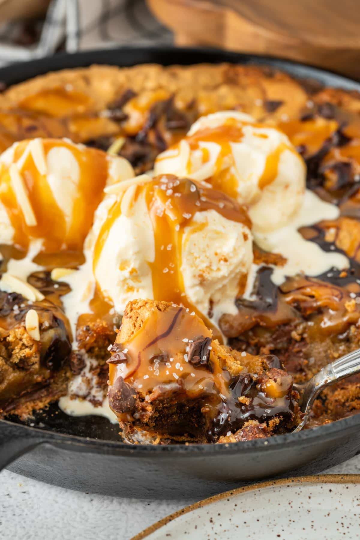 Molten caramel, melted chocolate, and melting ice cream turn this salted caramel chocolate chip skillet cookie into full dessert theater. Spoon lifting a gooey bite of salted caramel chocolate chip skillet cookie with molten caramel, melted chocolate, vanilla ice cream, and flaky sea salt in a cast iron skillet.