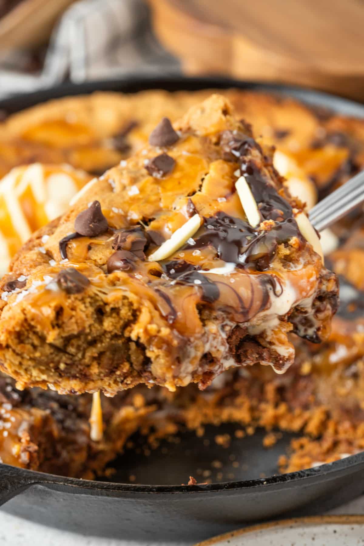 The molten caramel layer underneath keeps the center soft while the cast iron skillet creates crisp golden edges around the outside. Slice of salted caramel cookie skillet lifted from a cast iron pan with melted chocolate, caramel drizzle, flaky sea salt, and melting vanilla ice cream.