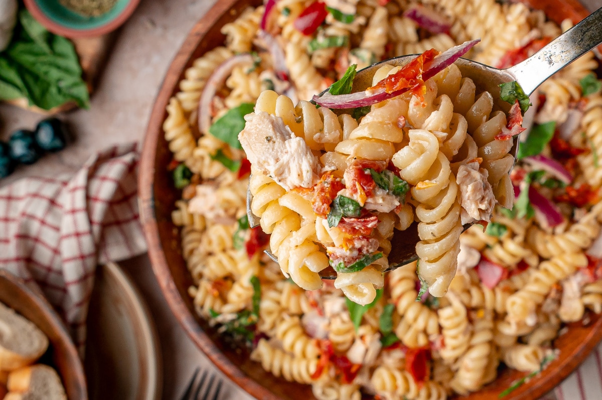 Spoonful of chicken pasta salad with basil, Parmesan, sun-dried tomatoes, and lightly coated rotini pasta Spoonful of Sun-Dried Tomato Chicken Pasta Salad with rotini, rotisserie chicken, basil, and red onion over a serving bowl