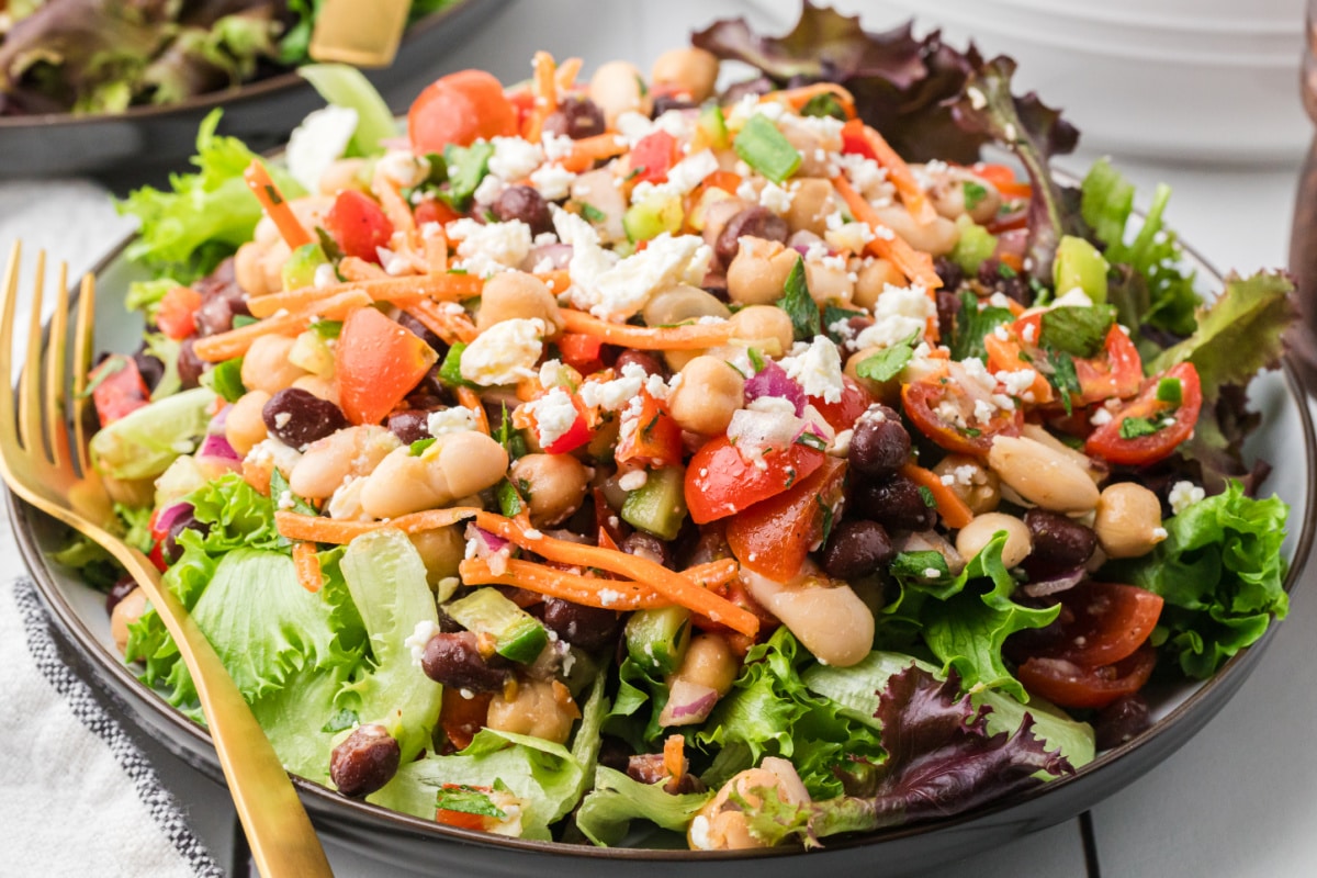 Three bean salad with feta cheese, chickpeas, black beans, cannellini beans, tomatoes, carrots, and cucumber over lettuce greens