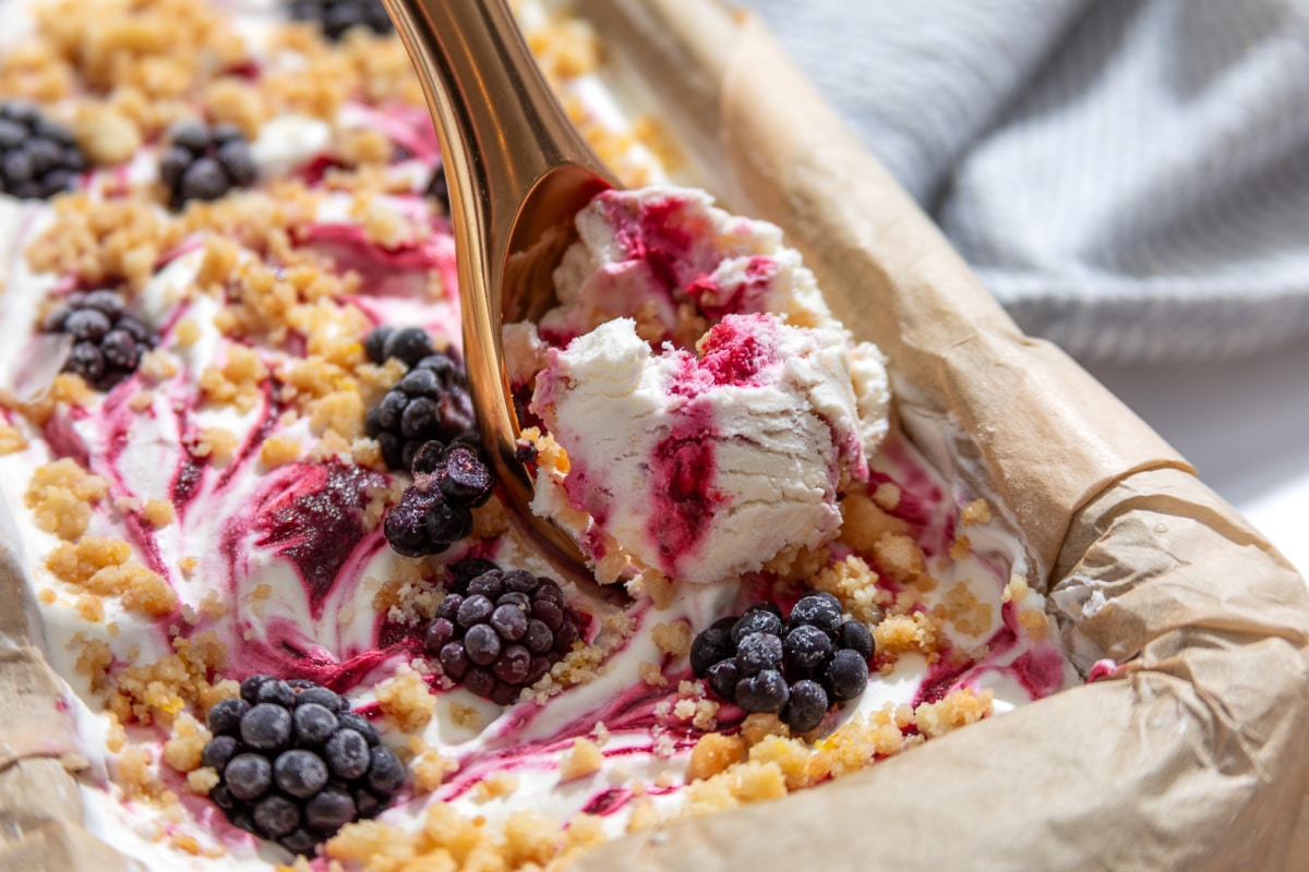 A scoop of Blackberry Crumble à la Mode Ice Cream with thick blackberry ribbons and buttery shortbread crumble being lifted from the loaf pan.