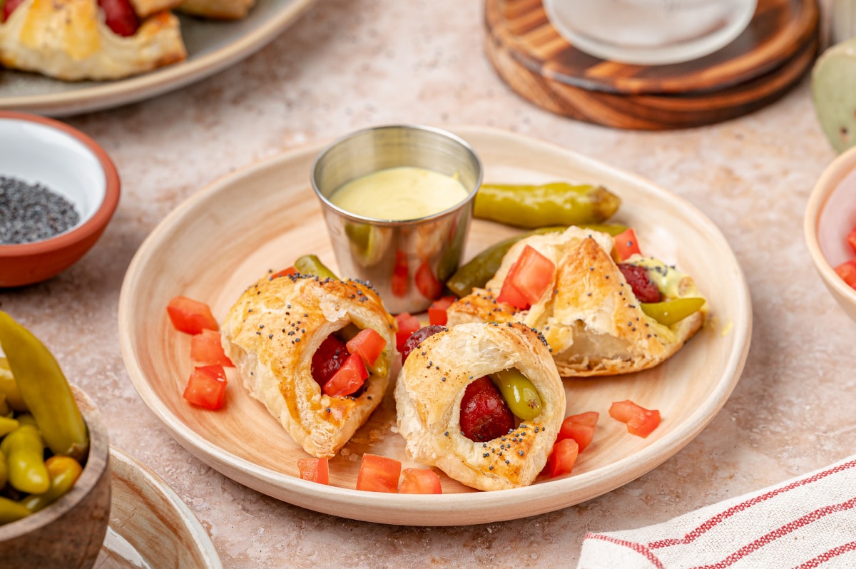 Close-up of Chicago hot dog pigs in a blanket with sport peppers, diced tomato, and creamy mustard relish dip.