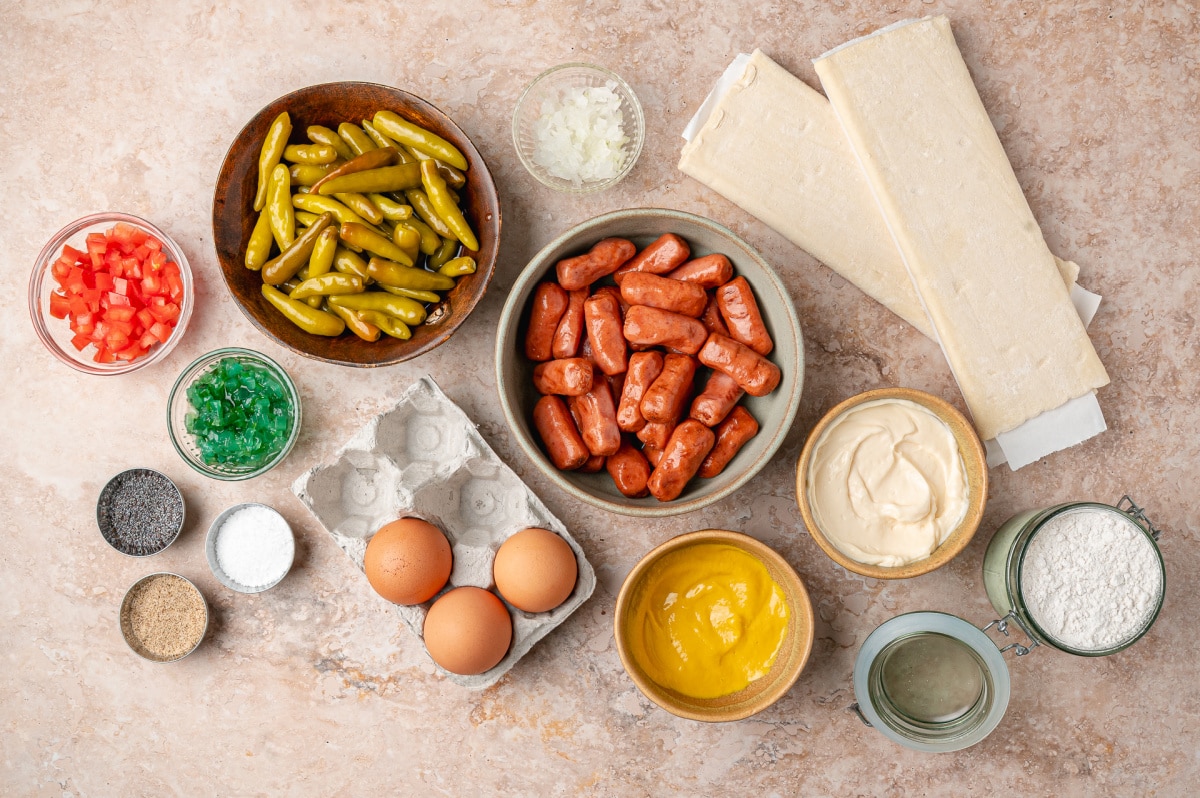 Ingredients for Chicago hot dog pigs in a blanket including puff pastry, beef cocktail franks, sport peppers, mustard, mayonnaise, relish, onion, tomato, and poppy seeds.