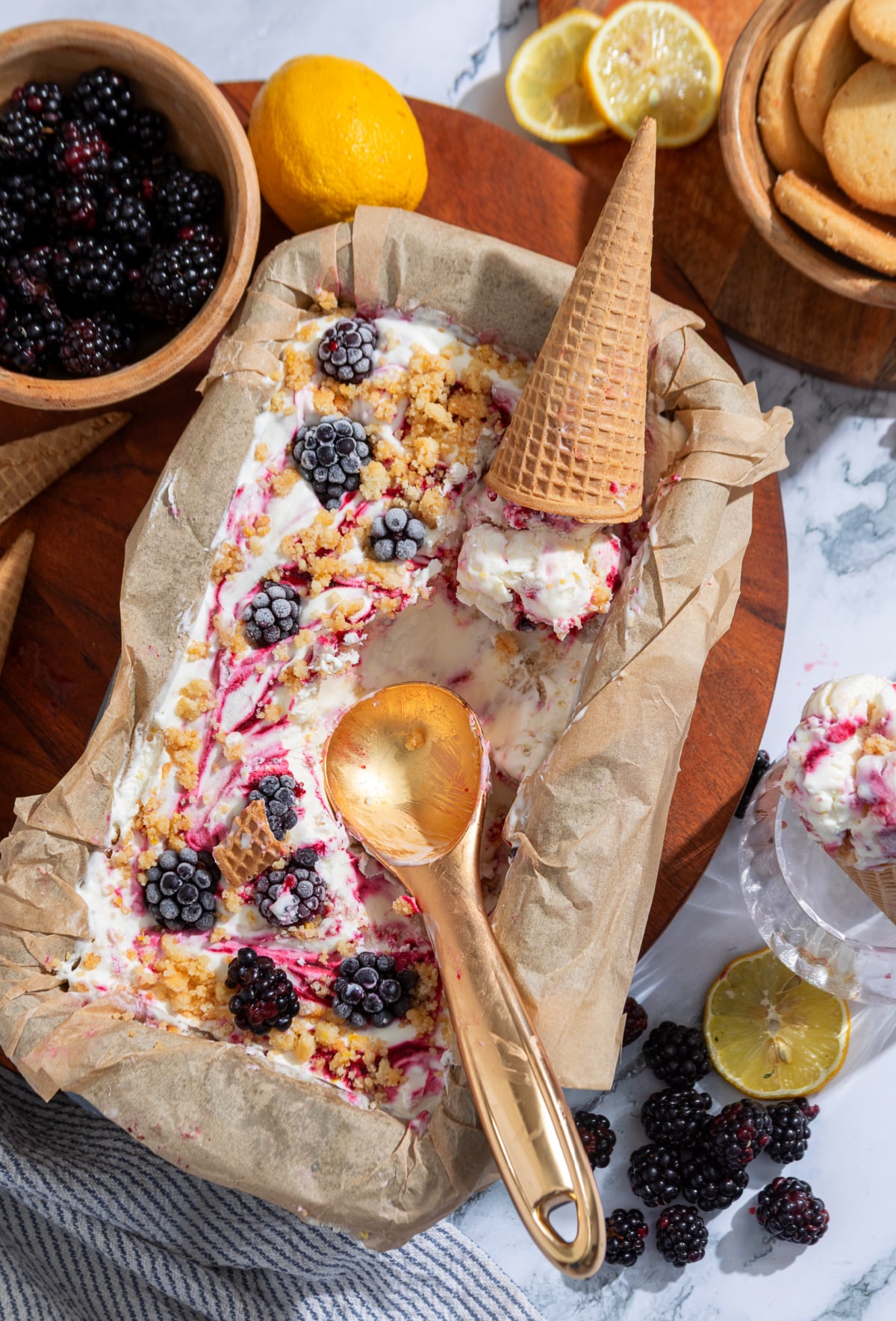 A loaf pan of homemade no-churn blackberry ice cream with blackberry swirls, shortbread crumble, fresh blackberries, and waffle cones.