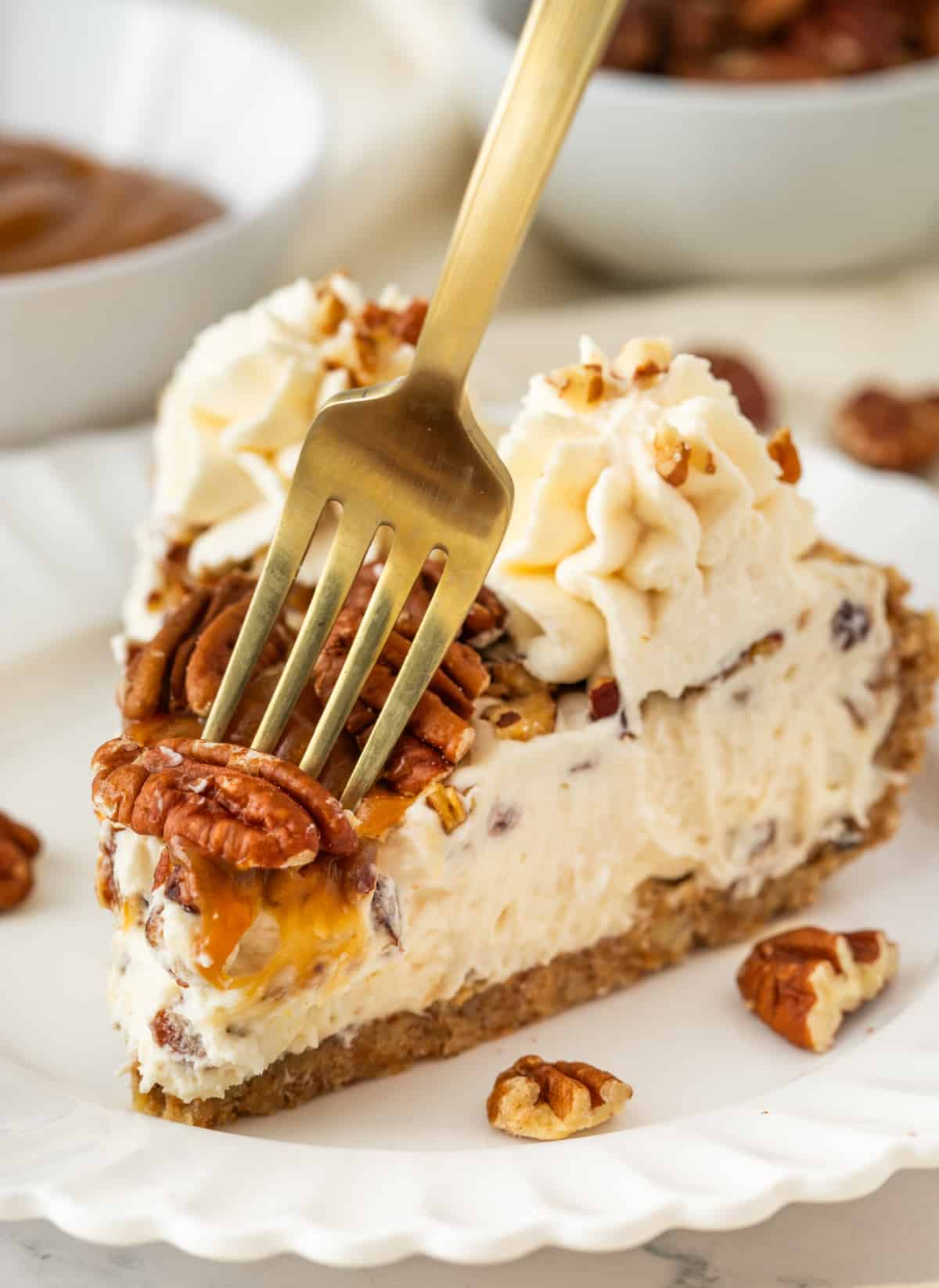 Fork pressing into a slice of pecan cream pie with toasted pecans, caramel topping, whipped cream, and pecan crust