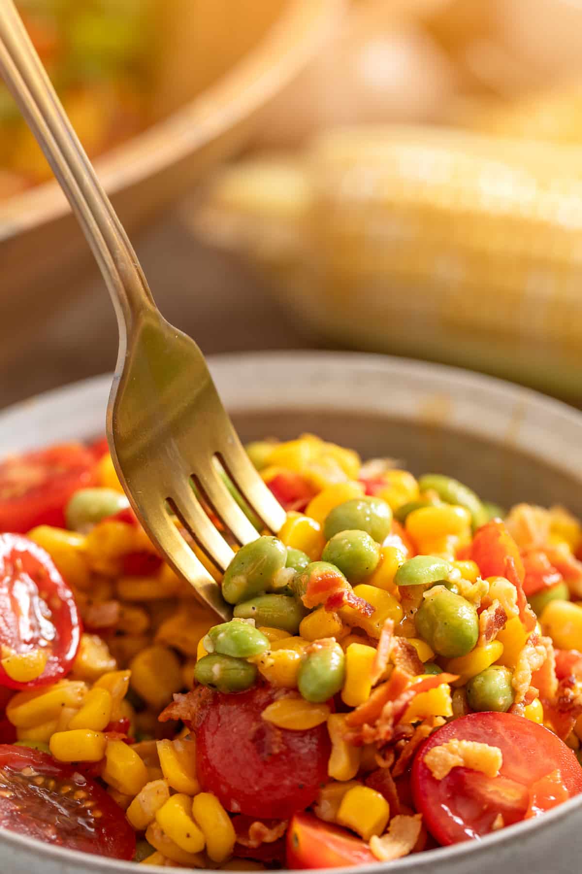 Fork lifting sweet corn, edamame, bacon, and tomatoes from a summer vegetable side dish