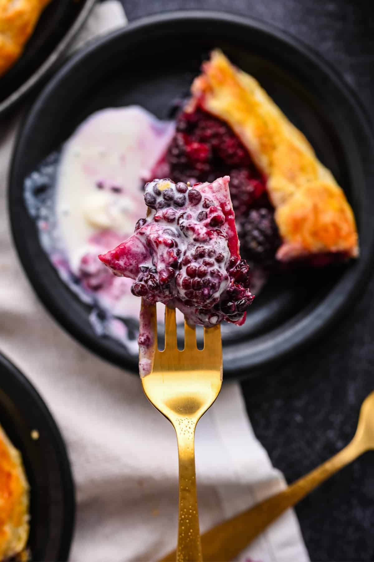 Fork holding a bite of blackberry puff pastry galette with melting vanilla ice cream.