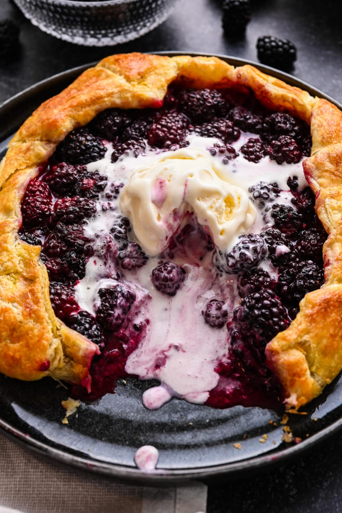 Blackberry puff pastry galette topped with vanilla ice cream on a black plate.