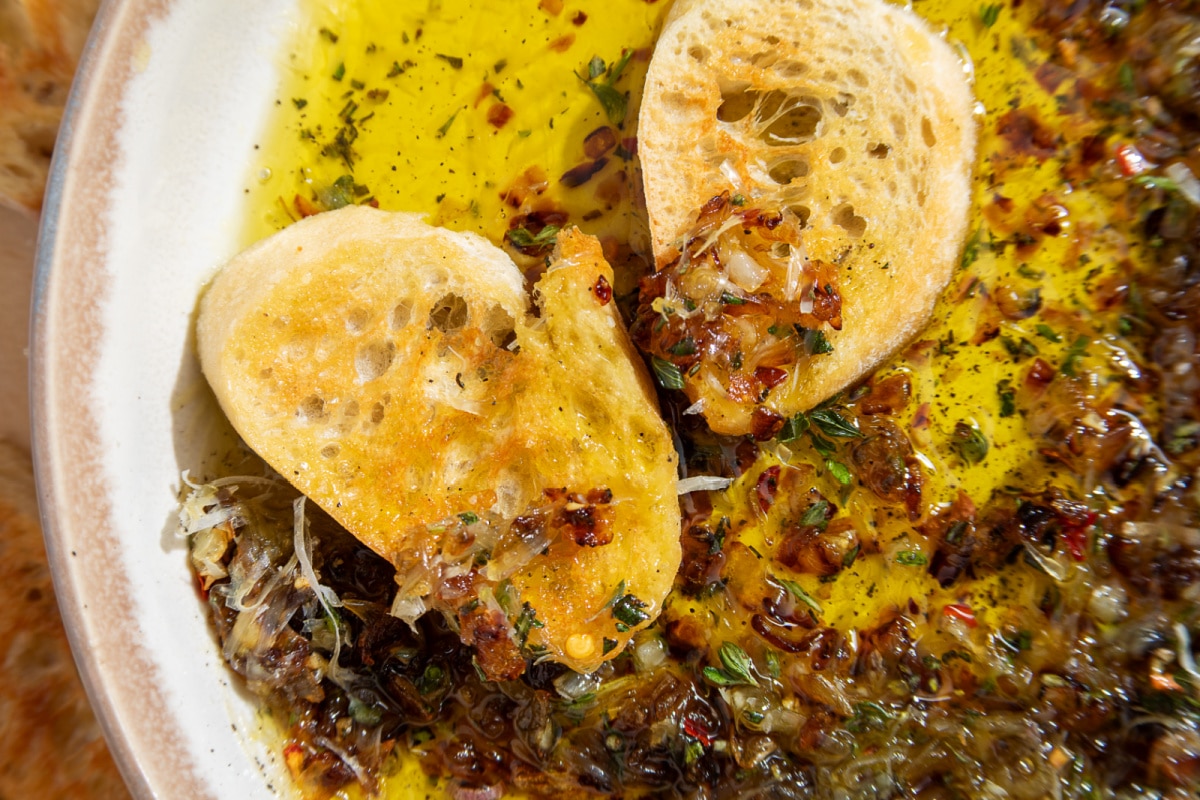 Close-up of toasted bread scooping caramelized shallots from bread dipping oil with fresh herbs and Parmesan.