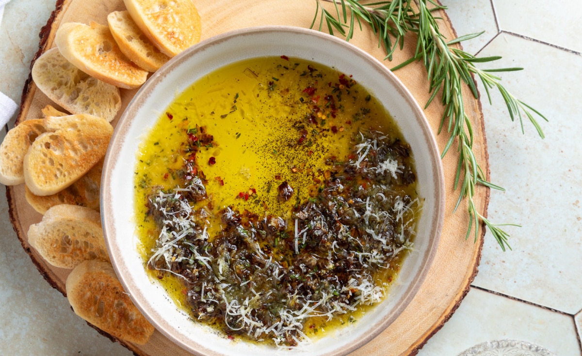 Overhead view of bread dipping oil with caramelized shallots, fresh herbs, Parmesan, and toasted baguette slices.