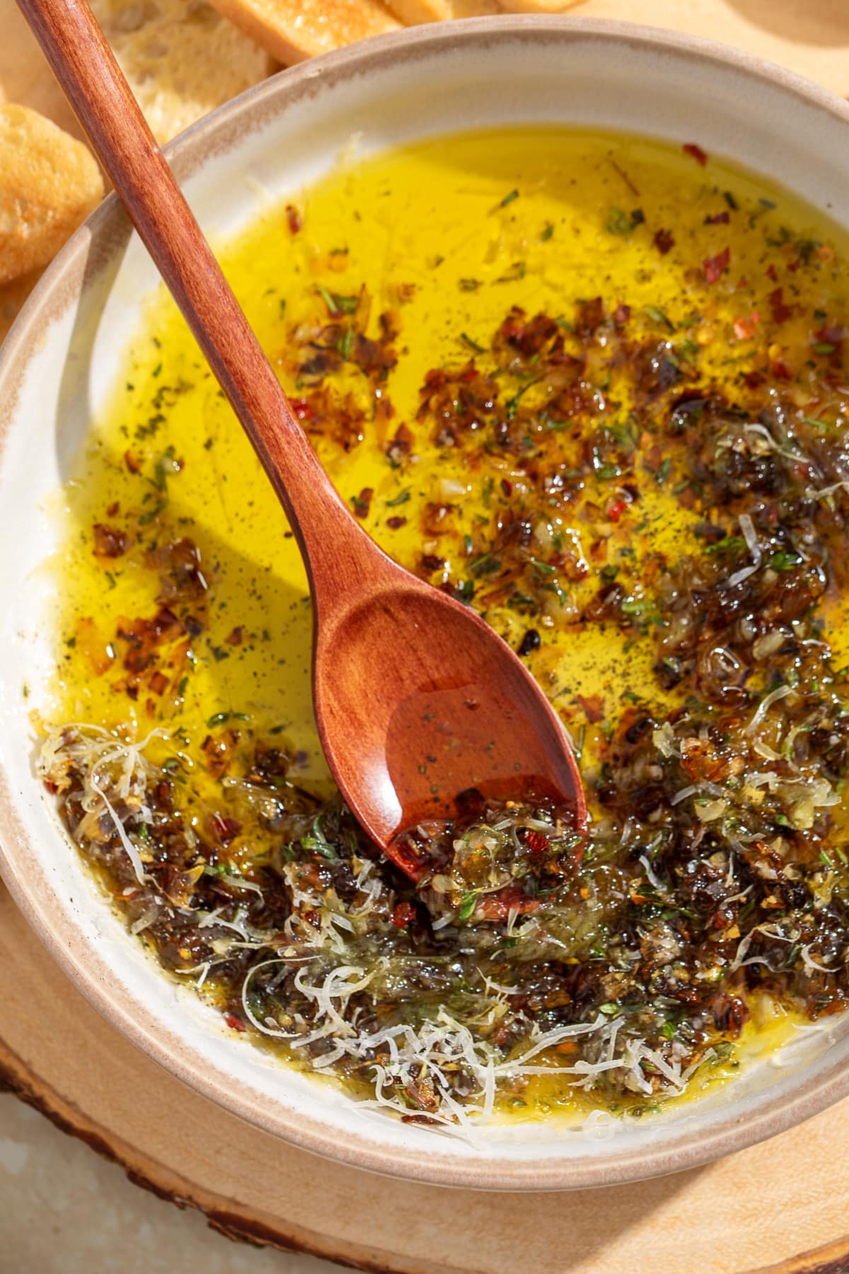 Wooden spoon scooping caramelized shallots from bread dipping oil with Parmesan, fresh herbs, and olive oil.