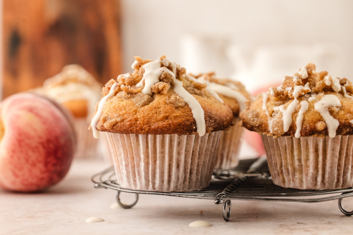 Brown butter peach coffee cake muffins topped with cinnamon streusel and vanilla glaze Brown butter peach coffee cake muffins cooling on a wire rack with fresh peaches