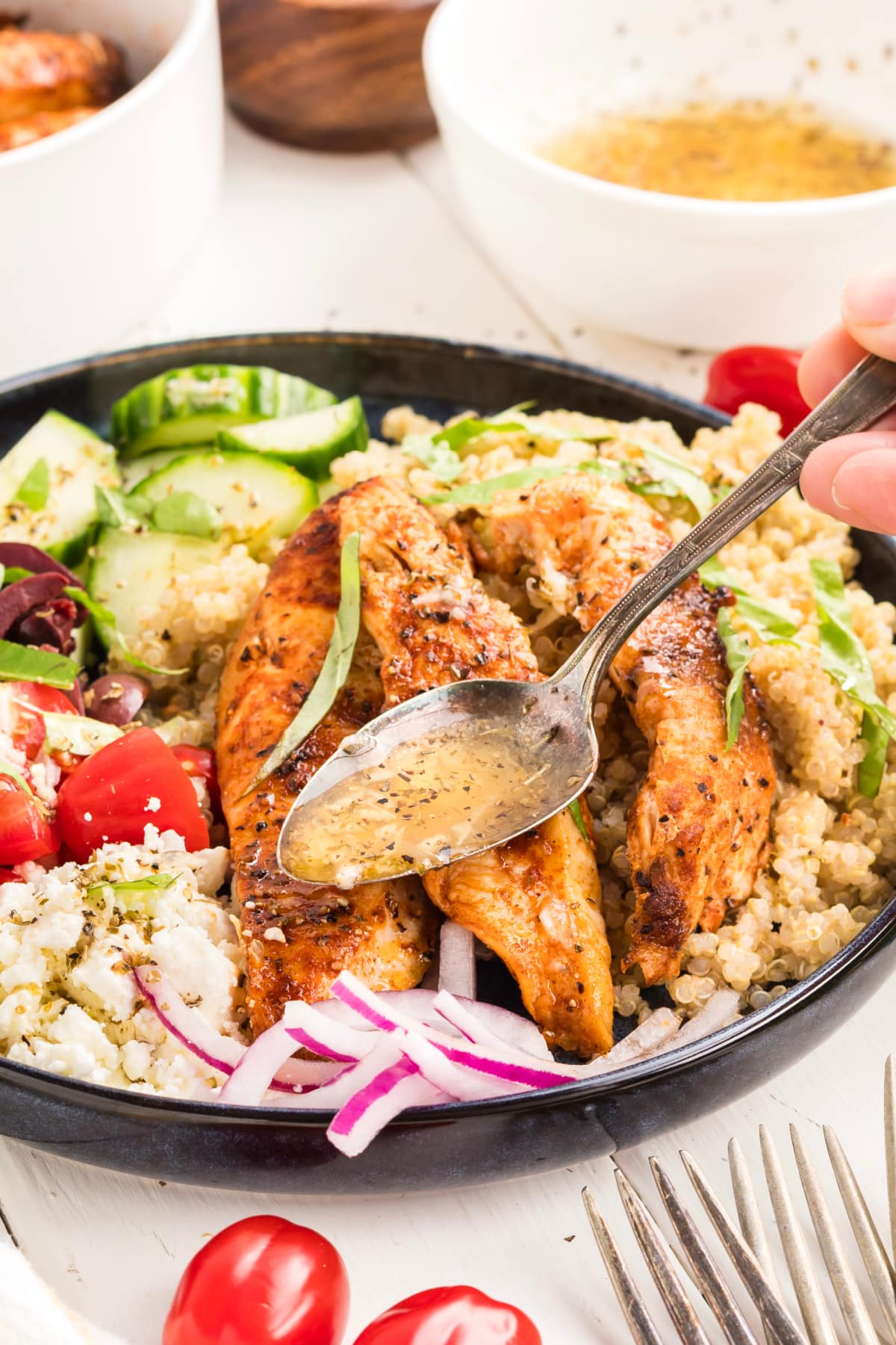 Spoon drizzling homemade lemon vinaigrette over a Mediterranean chicken bowl with grilled chicken, quinoa, feta, tomatoes, cucumber, olives, and red onion.