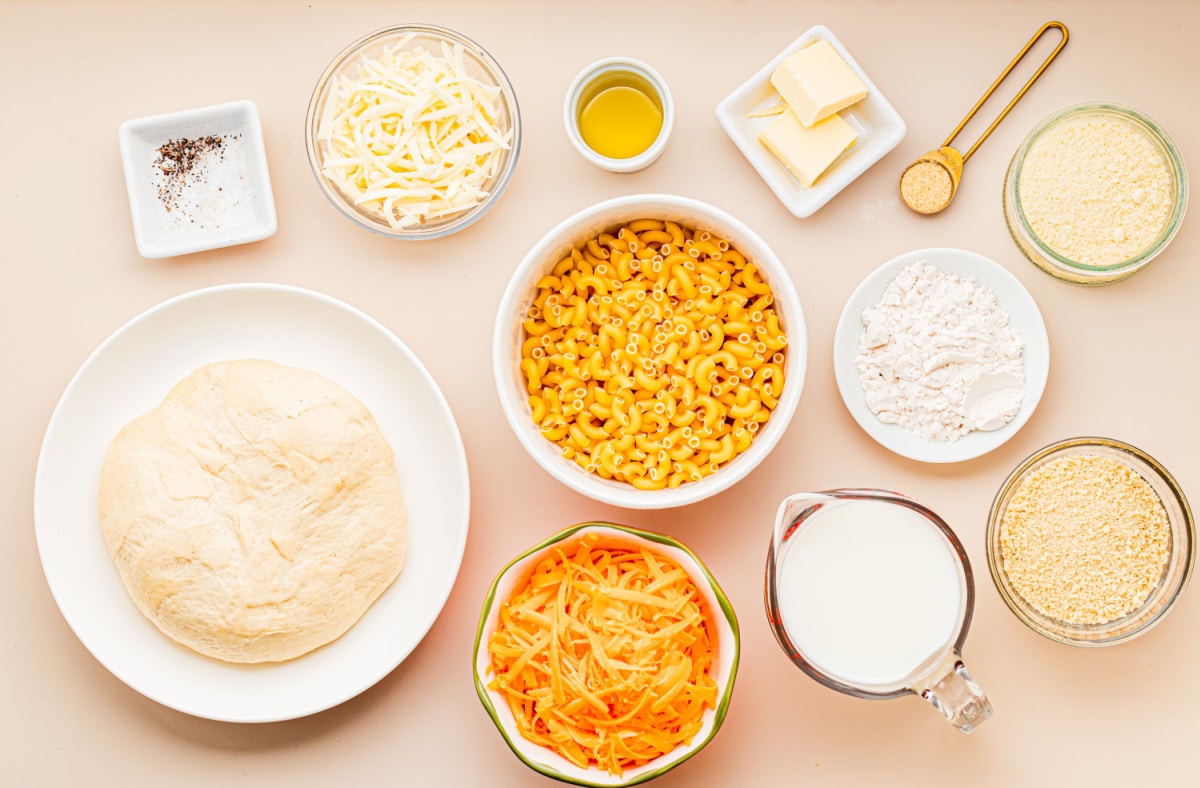 Ingredients for homemade mac and cheese pizza including pizza dough, elbow macaroni, cheddar, mozzarella, Parmesan, milk, butter, flour, olive oil, and panko breadcrumbs.