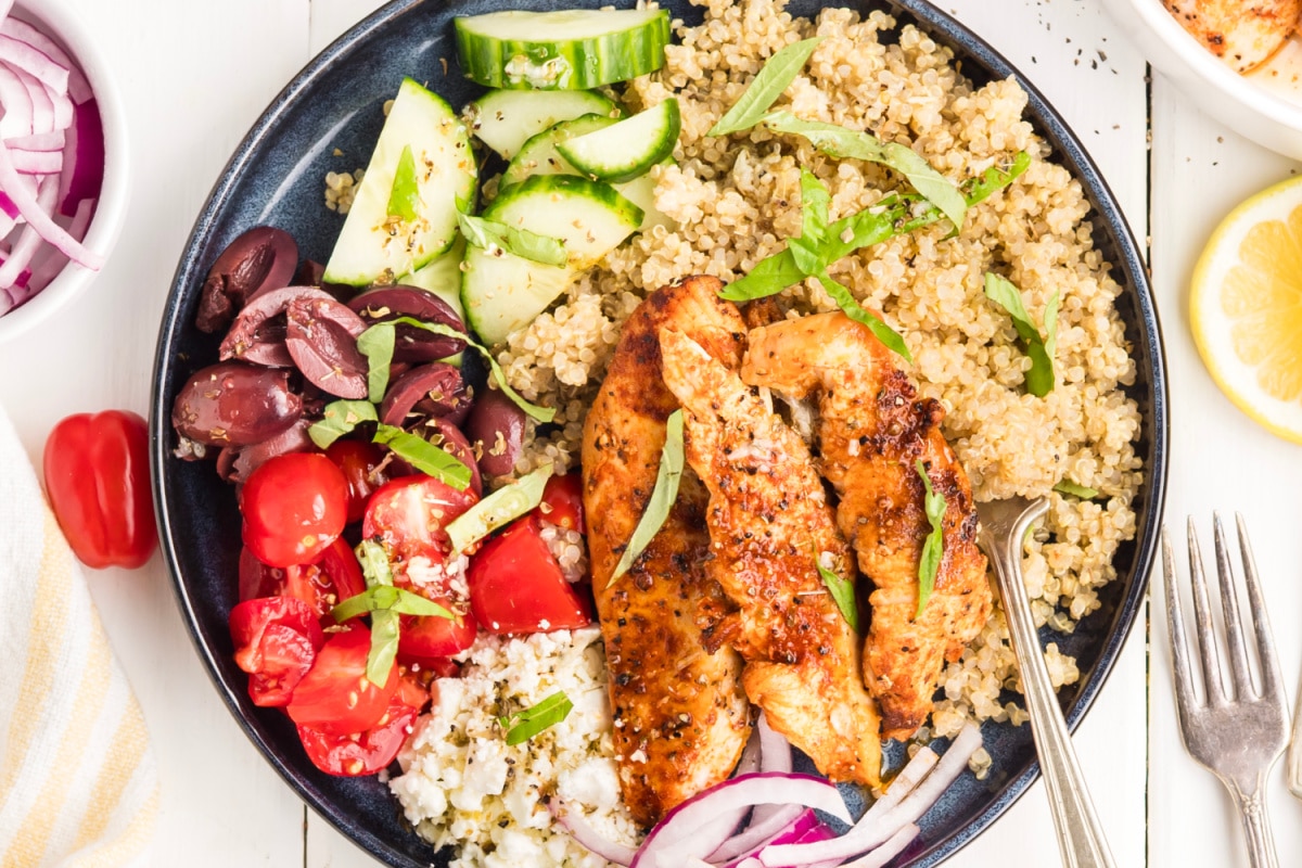 Overhead view of a Mediterranean chicken bowl with grilled chicken, quinoa, cherry tomatoes, cucumber, feta, Kalamata olives, red onion, basil, and lemon vinaigrette.