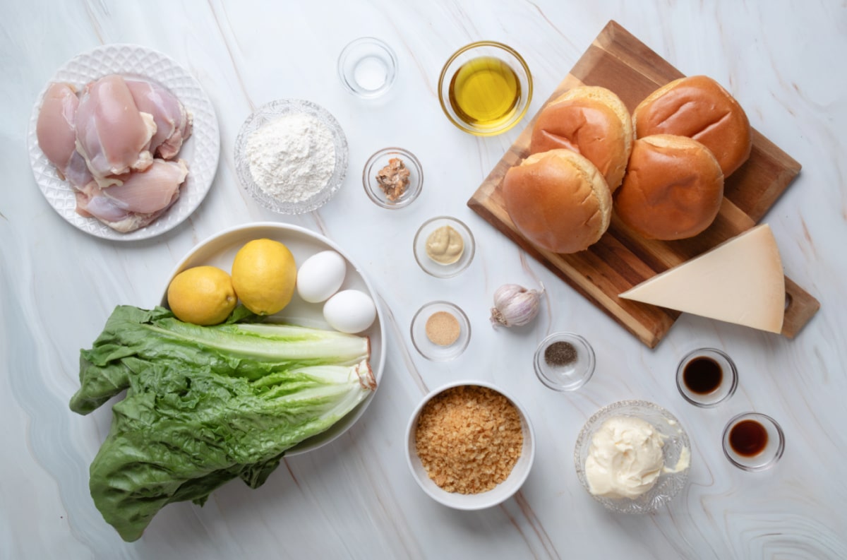 Ingredients for a chicken Caesar salad sandwich, including chicken thighs, romaine lettuce, Parmesan cheese, potato chips, sandwich rolls, eggs, lemons, olive oil, and Caesar dressing ingredients.