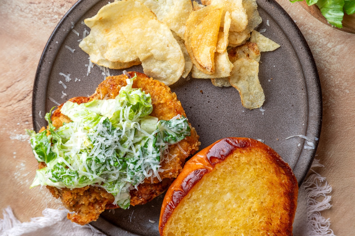 Overhead view of a chicken Caesar salad sandwich with crispy potato chip-crusted chicken, homemade Caesar salad, toasted brioche bun, and potato chips.
