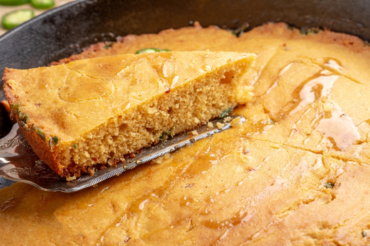 Slice of chipotle jalapeño cornbread lifted from a cast-iron skillet with hot honey drizzled over the top