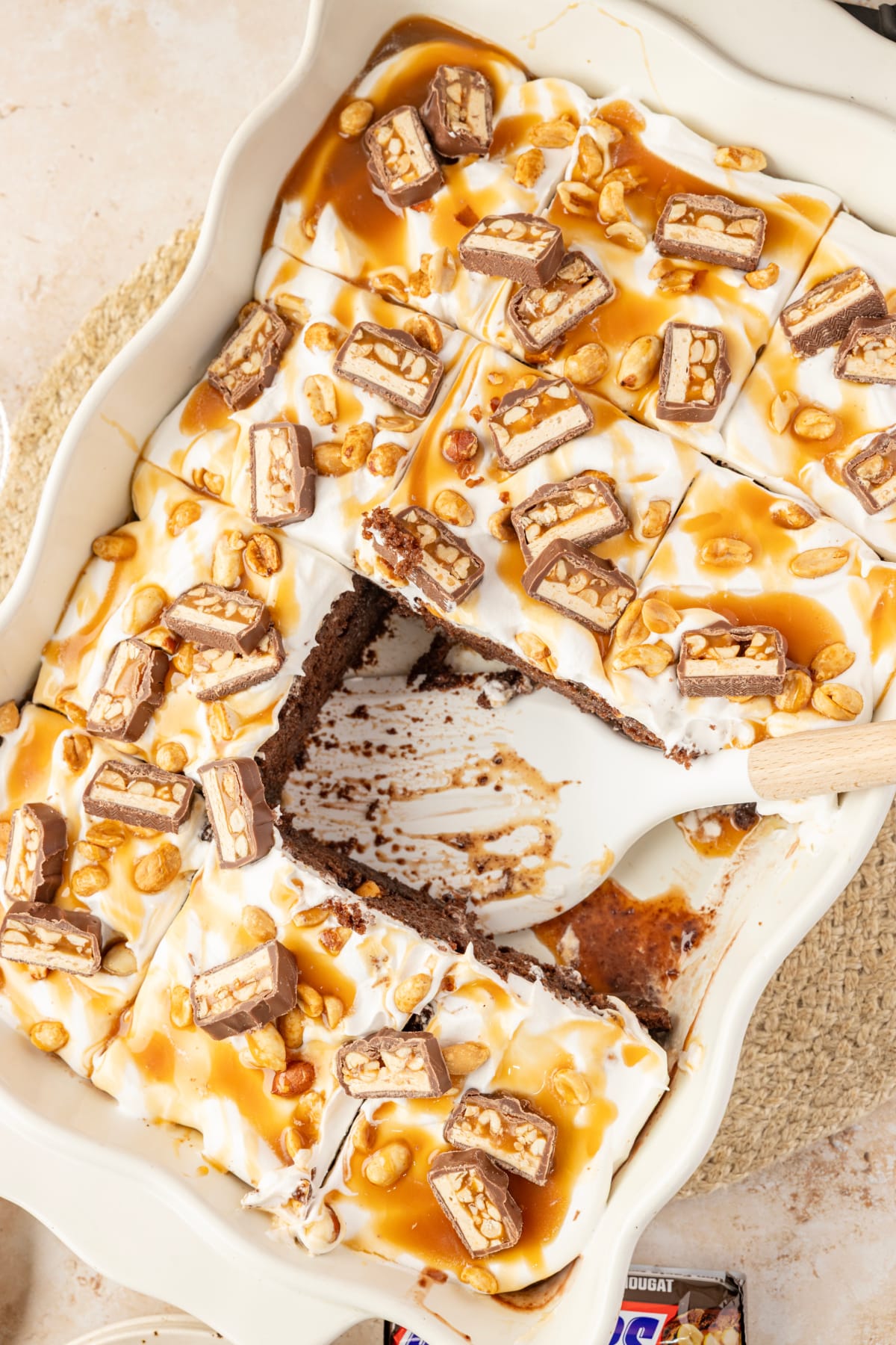 Chocolate pudding poke cake cut into squares with caramel, salted peanuts and chopped Snickers bars