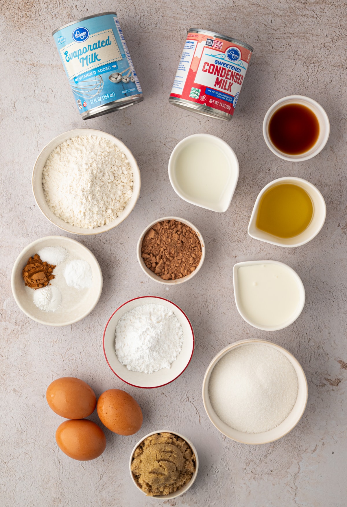 Ingredients for chocolate tres leches cake including cocoa powder, three milks, eggs, flour, sugar, oil, cinnamon, and vanilla.