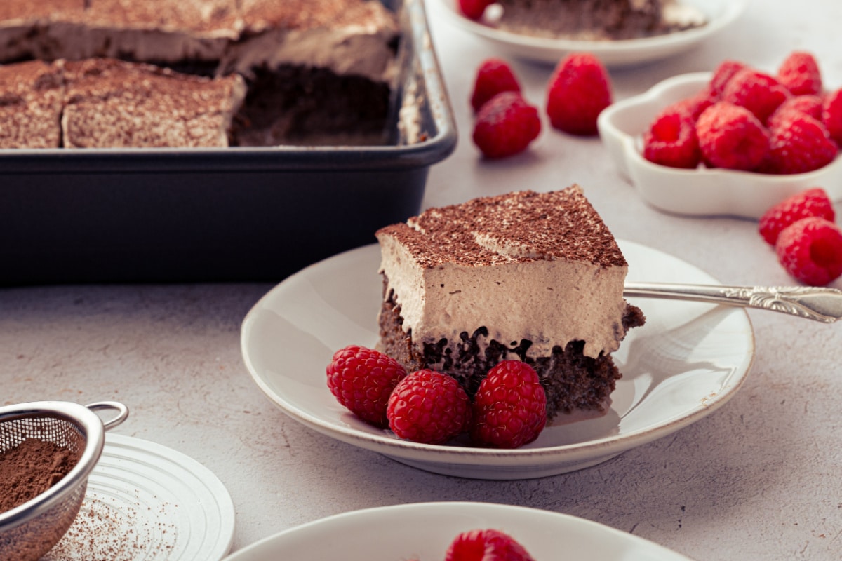 Slice of chocolate tres leches cake with chocolate whipped cream and raspberries beside the baking pan.