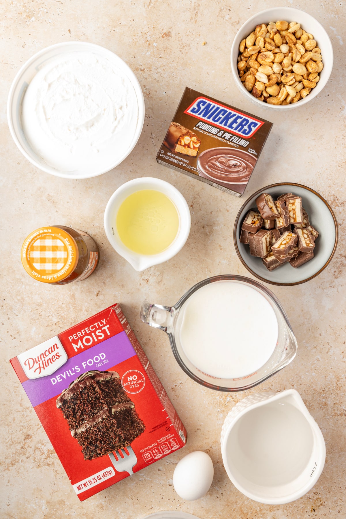 Ingredients for Snickers poke cake with chocolate cake mix, pudding, caramel, peanuts and chopped Snickers bars