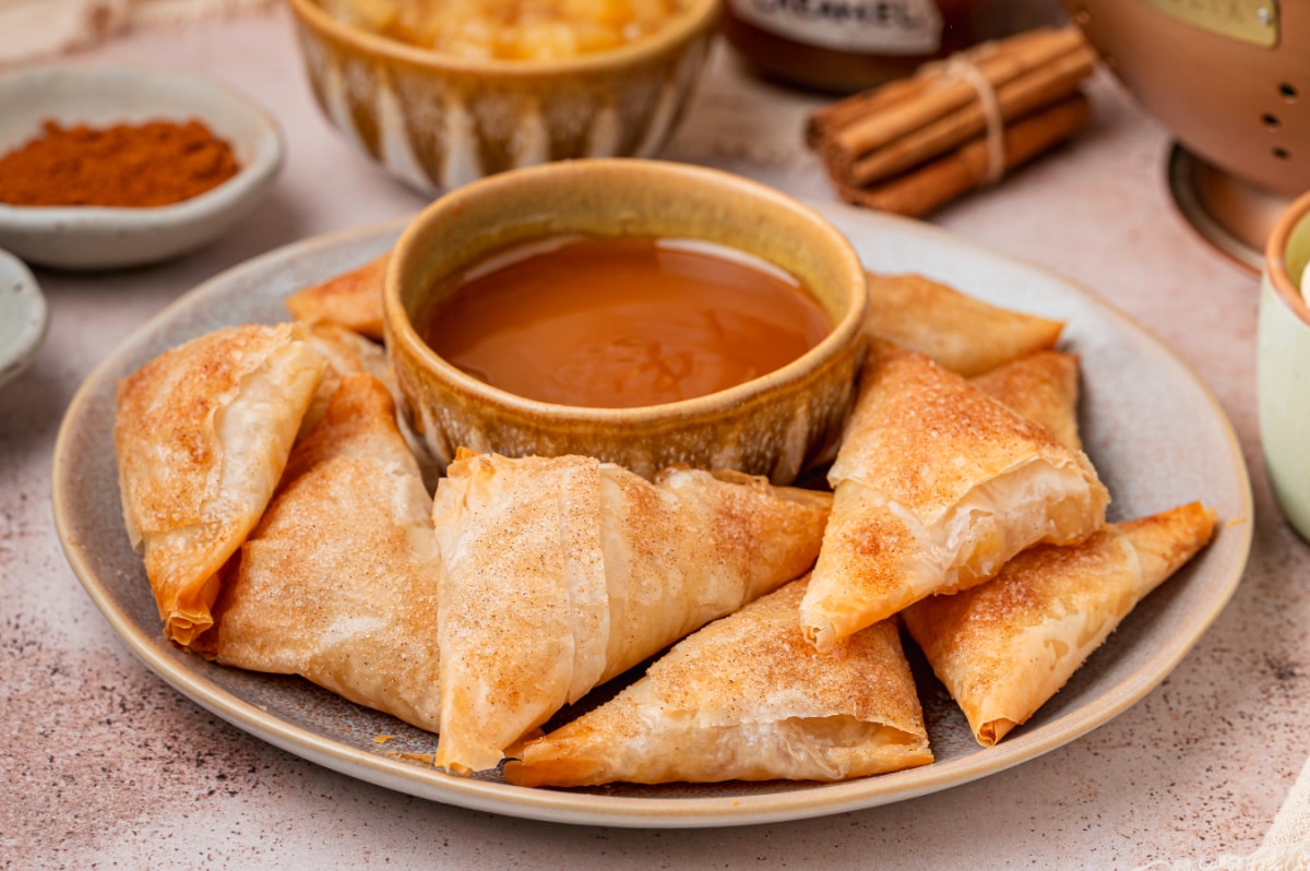 Cinnamon sugar apple pie samosas arranged on a plate with caramel sauce for dipping.