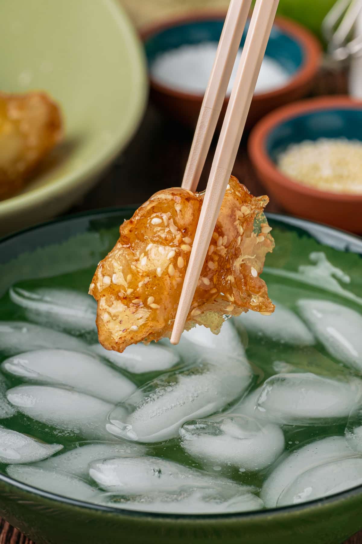 Caramel-coated fried apple held with chopsticks over a bowl of ice water before dipping