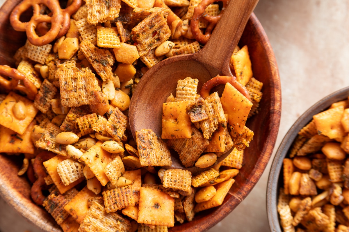 Herbed cowboy butter snack mix with a wooden spoon showing the Chex, crackers and nuts. Cowboy butter snack mix with Chex cereal, Cheez-It crackers, Triscuits, pretzels, peanuts and walnuts in a wooden bowl
