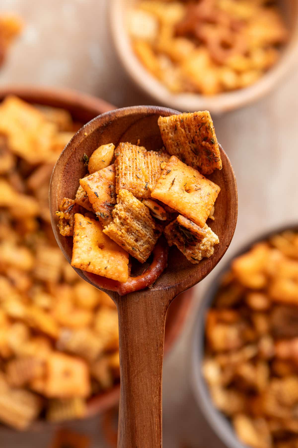 Close-up of the different cereals and crackers after baking with the herbed butter mixture. Wooden spoon filled with seasoned Chex cereal, Cheez-It crackers, Triscuit pieces and a pretzel