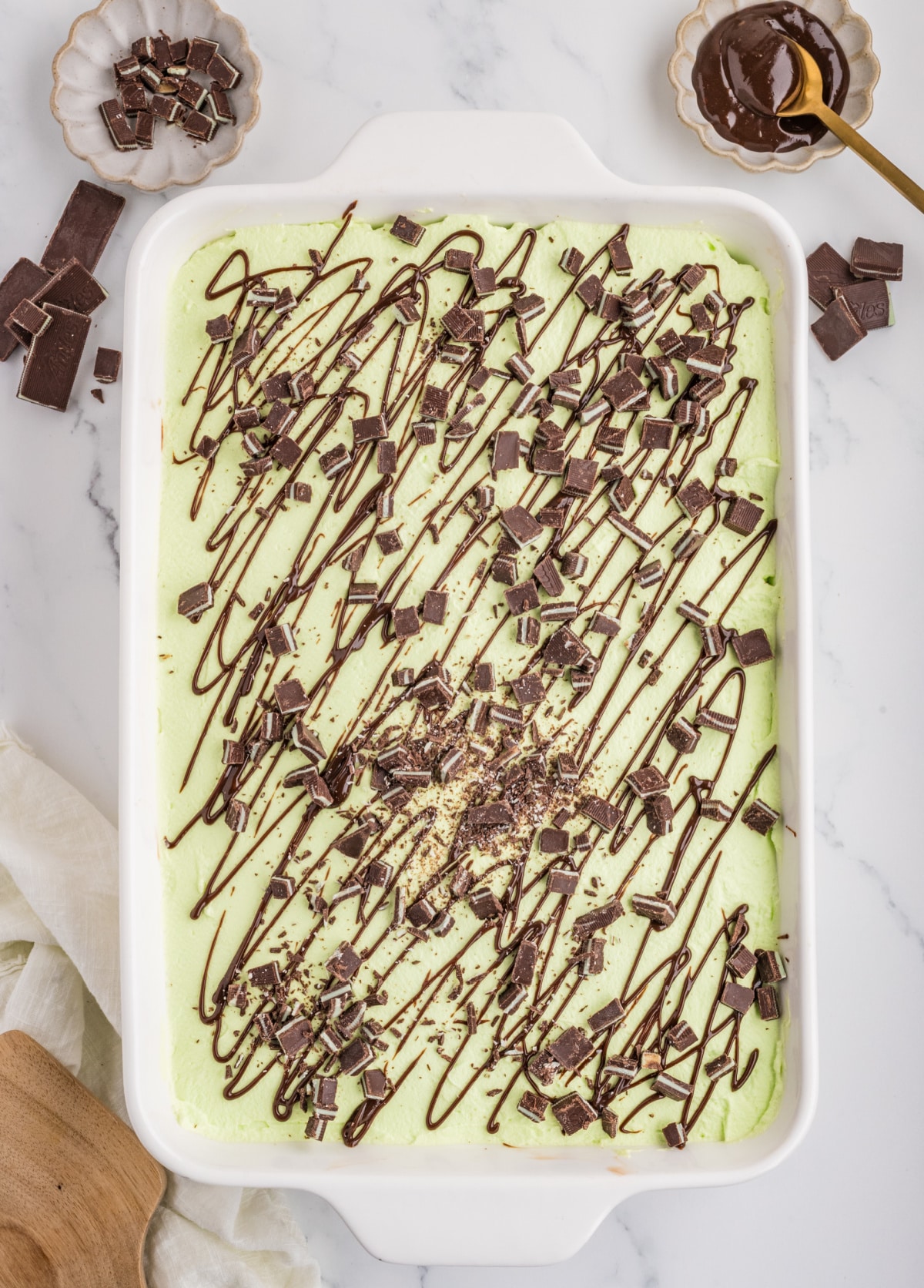 Finished chocolate poke cake in a 9×13-inch baking dish topped with pale green mint whipped cream, chopped Andes mints and hot fudge drizzle.