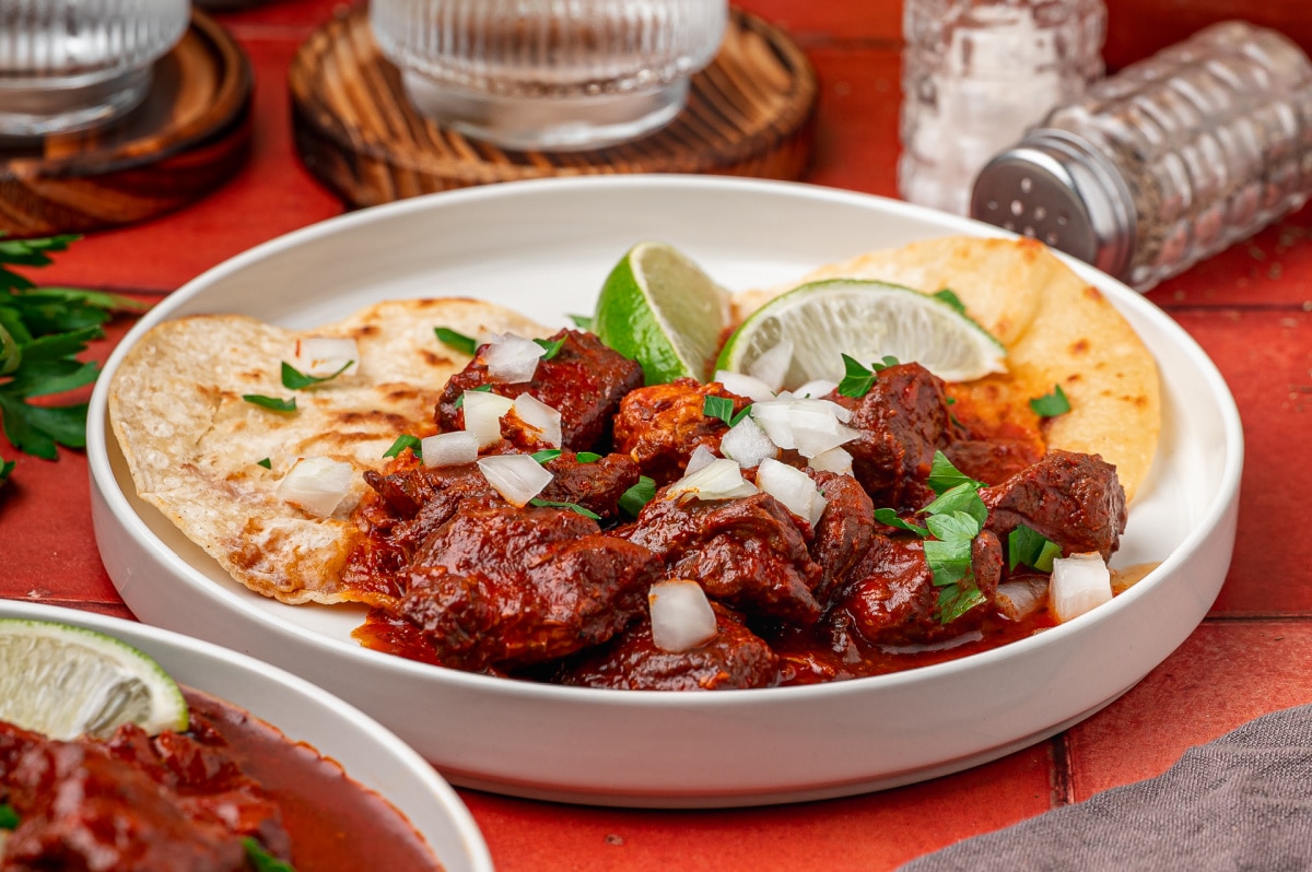 Fork holding tender pork from carne adovada in a red chile sauce Carne adovada served with warm tortillas, lime, chopped onion and parsley
