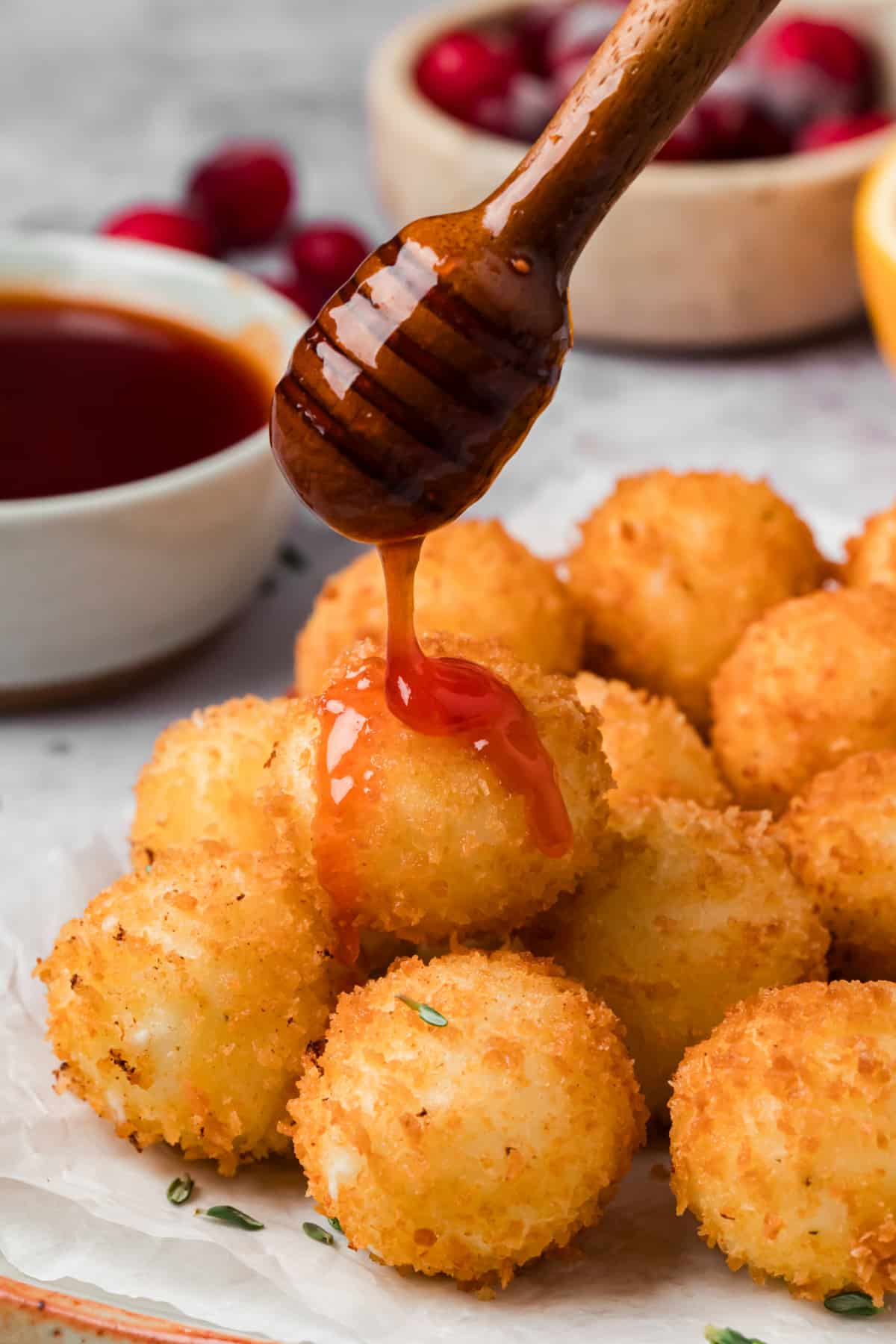 Cranberry hot honey dripping from a wooden honey dipper onto a golden panko-crusted goat cheese ball.