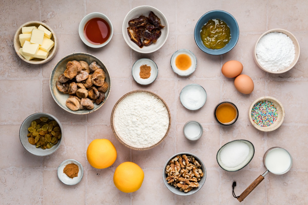 Ingredients for Homemade Italian Fig Cookies (Cuccidati) Ingredients for homemade cuccidati arranged in bowls, including dried figs, dates, golden raisins, walnuts, oranges, flour, butter, eggs, and glaze ingredients.