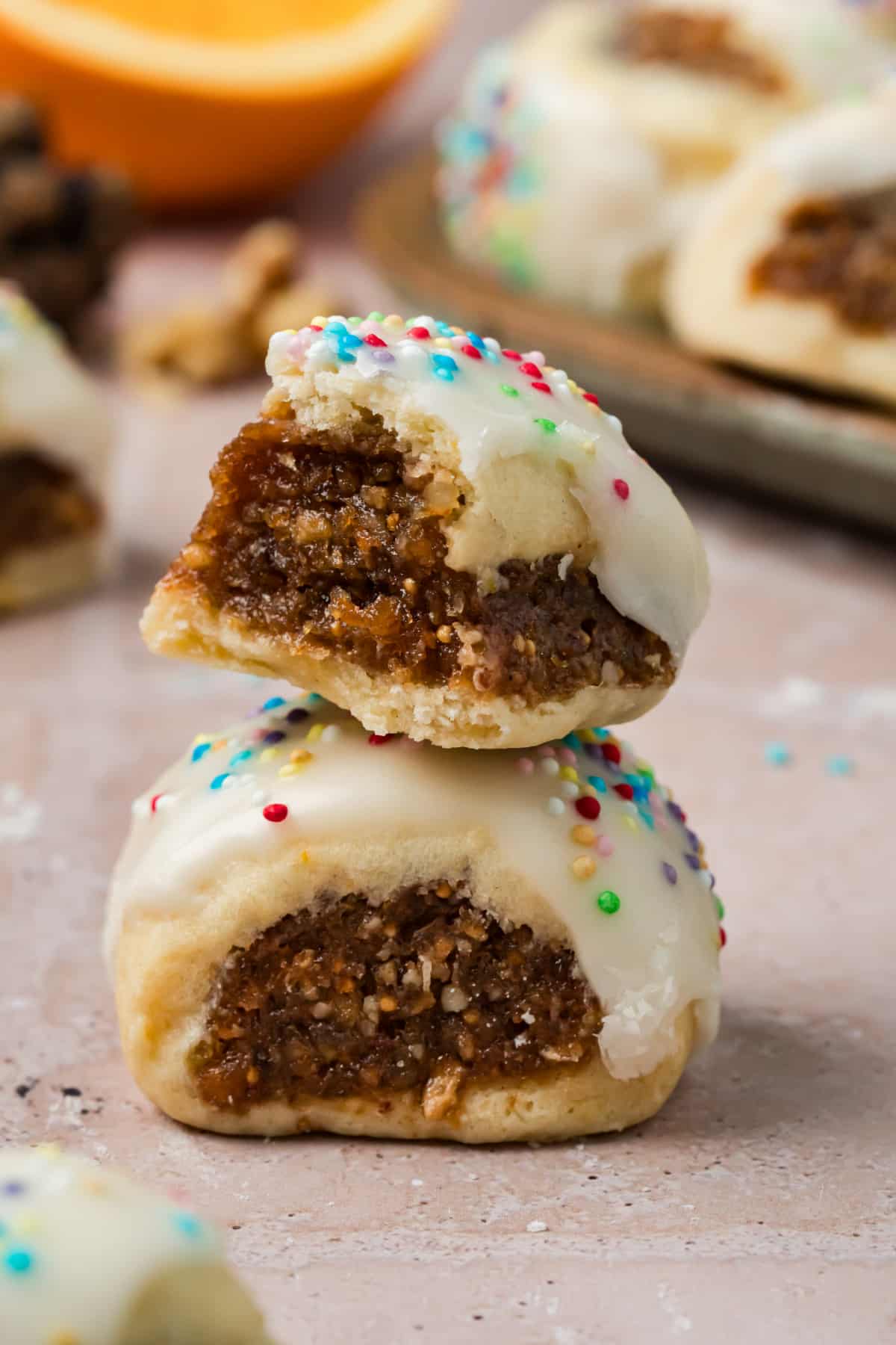 Cuccidati Cookies with Moist Fig Filling and Vanilla Glaze Close-up of a bitten cuccidati cookie stacked on another, showing the moist fig, date, and walnut filling, tender pastry, vanilla glaze, and rainbow sprinkles.