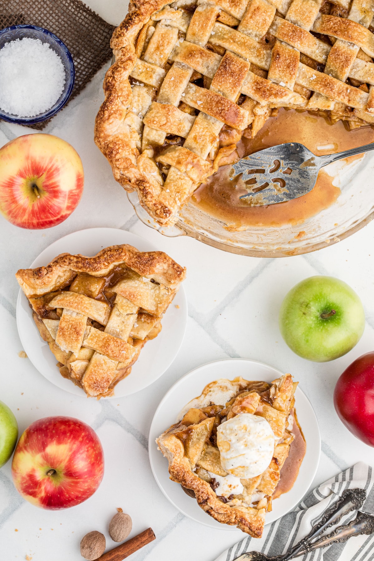 Overhead view of homemade caramel apple pie with a slice removed, two plated slices, vanilla ice cream, and caramel sauce pooled in the pie plate.