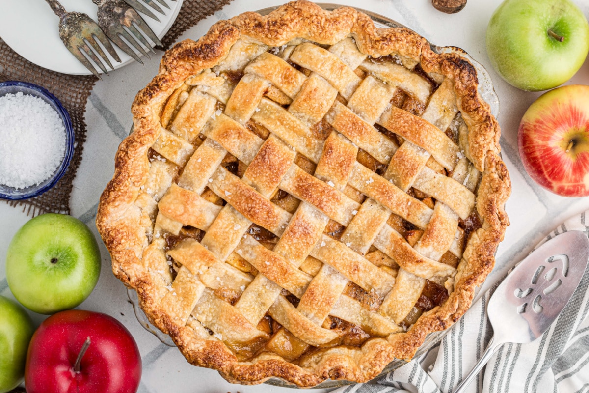 Overhead view of a homemade caramel apple pie with a golden all-butter lattice crust sprinkled with flaky sea salt, surrounded by fresh apples.