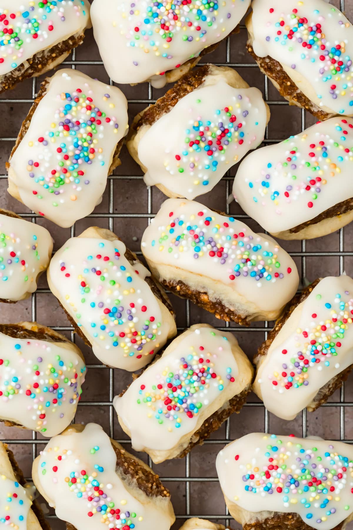 Italian Fig Cookies with Vanilla Icing and Colorful Nonpareils Overhead close-up of Italian fig cookies on a wire rack, showing thick white vanilla glaze, rainbow nonpareils, and fig filling along the sides.