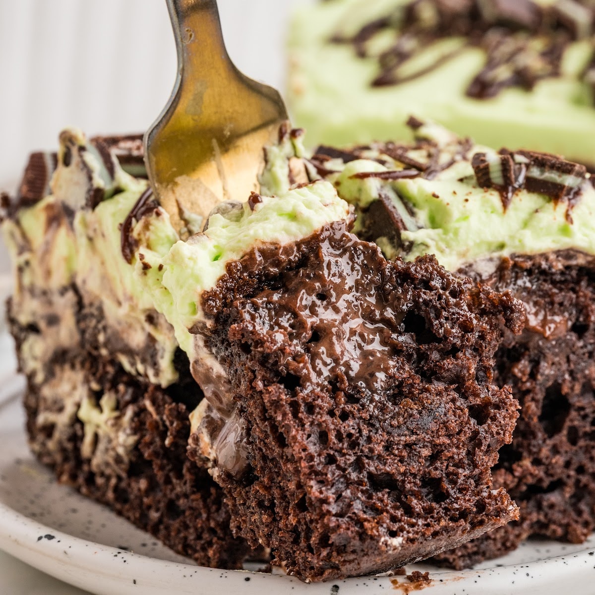 Close-up of mint chocolate poke cake with a fork lifting a piece of chocolate cake filled with hot fudge and topped with pale green mint whipped cream.