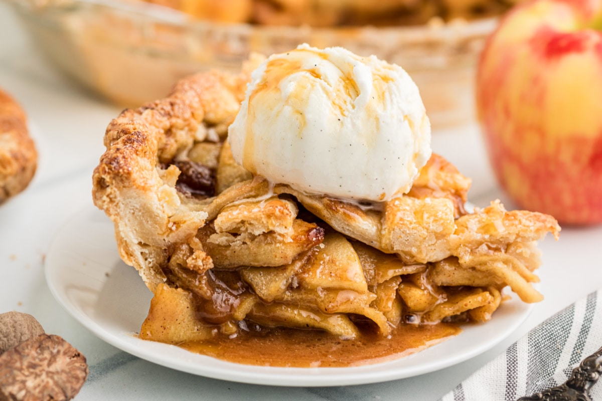 Close-up of salted caramel apple pie topped with vanilla ice cream, showing thin apple slices coated in caramel sauce beneath a flaky crust.